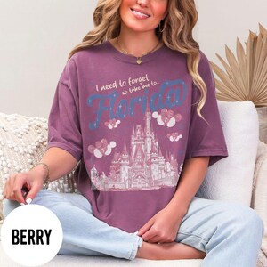 May include: Berry-colored t-shirt with the text "I need to forget so take me to Florida" in blue script. The design includes a castle illustration and decorative elements. The shirt is a comfortable, oversized fit.