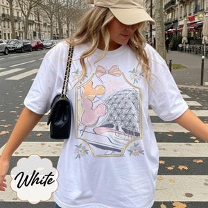 May include: White t-shirt featuring a colorful graphic design with a bow, Mickey Mouse balloons, and a geometric sphere. The shirt has short sleeves and a relaxed fit. The word "White" is written in a cursive font on the bottom left.
