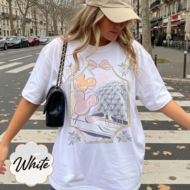 May include: White t-shirt featuring a colorful graphic design with a bow, Mickey Mouse balloons, and a geometric sphere. The shirt has short sleeves and a relaxed fit. The word "White" is written in a cursive font on the bottom left.