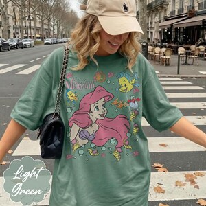May include: Light green t-shirt with a graphic of Ariel from Disney's The Little Mermaid. The design includes Ariel with pink hair, a purple seashell bra, and various sea creatures. The shirt also has the text "The Little Mermaid".