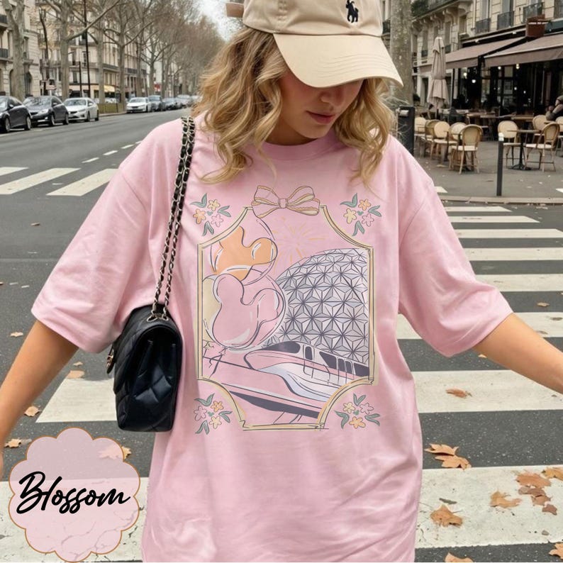 May include: A light pink t-shirt with a graphic featuring a cartoon character, a geometric sphere, and a train. The design is framed with floral and bow accents. The word "Blossom" is written in cursive at the bottom.