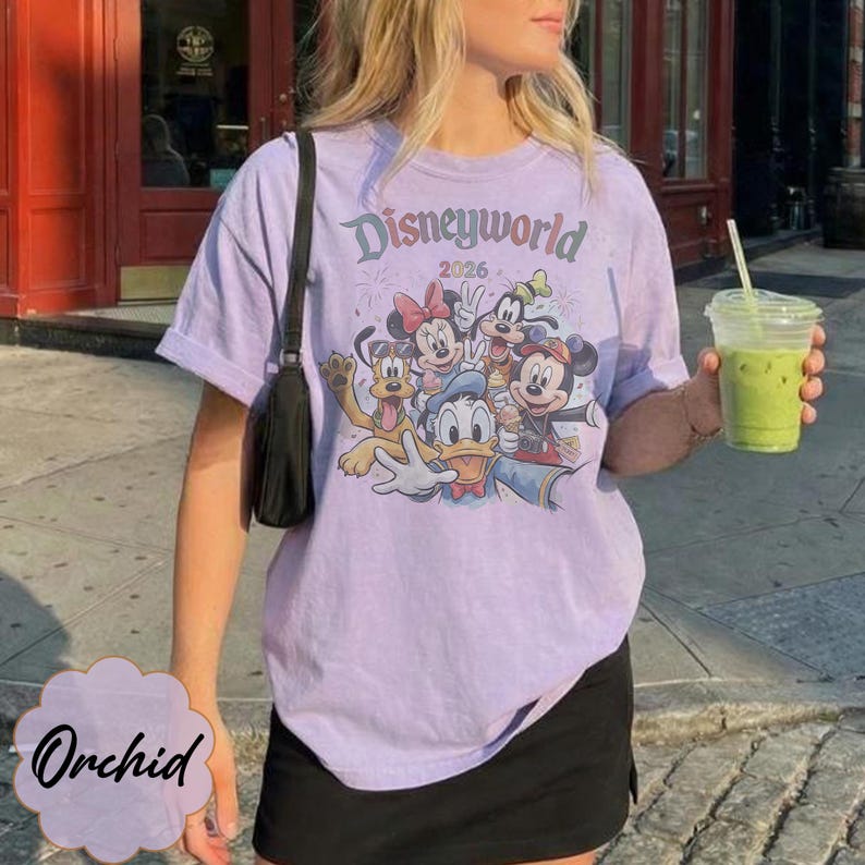 Pu&ograve; includere: Una maglietta lilla con la scritta "Disneyworld 2026" e una grafica dei personaggi Disney. La maglietta ha maniche corte e girocollo. Nell'immagine sono visibili anche una borsa a tracolla nera e una bevanda verde in un bicchiere trasparente.