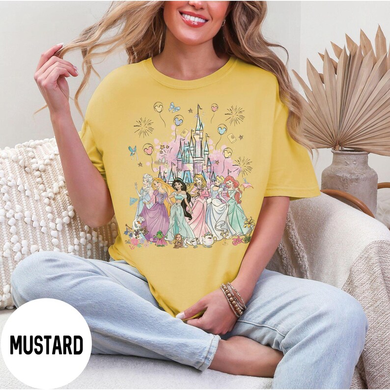 May include: Mustard-colored t-shirt featuring a colorful graphic of several princesses in front of a castle, with fireworks and balloons. The word "MUSTARD" is printed at the bottom. The shirt is a casual, comfortable style.
