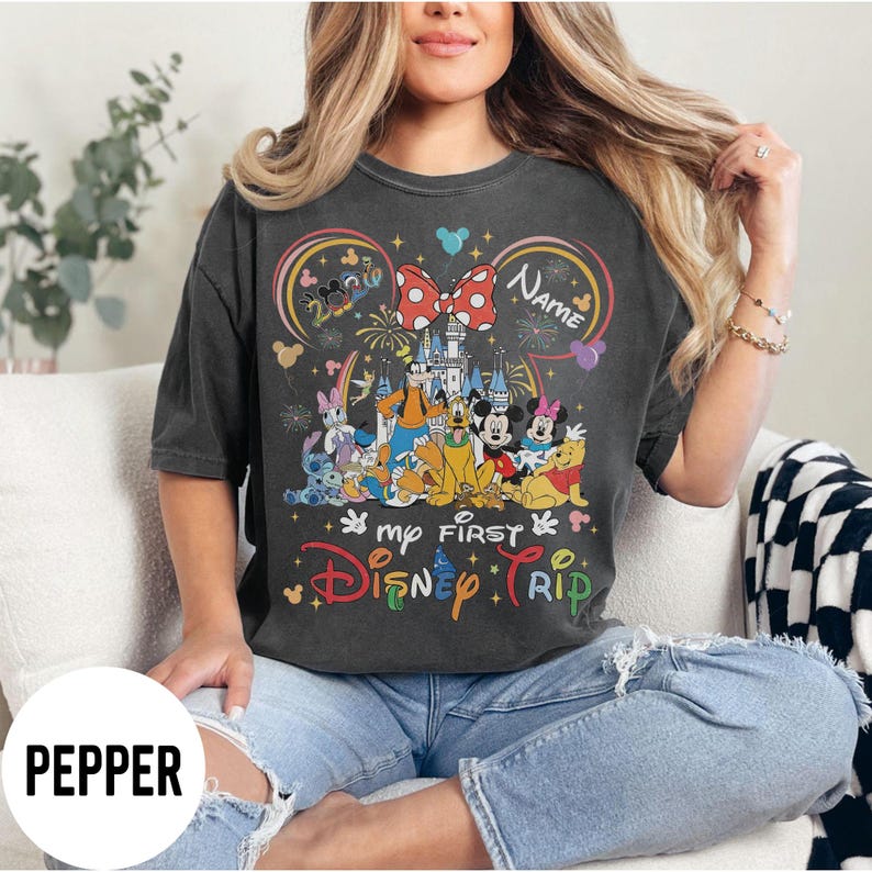 May include: Dark gray t-shirt featuring a colorful Disney design. The design includes various Disney characters, a castle, fireworks, and the text "my FIRST Disney TRIP". A red bow with white polka dots and the word "Name" are also featured.