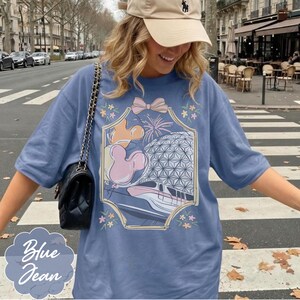 May include: A blue jean t-shirt featuring a framed graphic with a pink bow, Mickey Mouse balloons, fireworks, and a futuristic building. The shirt has short sleeves and a relaxed fit. The words "Blue Jean" are in a light blue oval at the bottom.