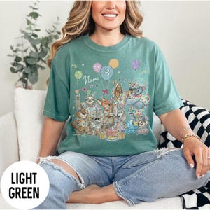 以下が含まれることがあります： 誕生日を祝う動物たちが描かれた、カラフルな漫画デザインのライトグリーンのTシャツ。デザインには風船、ケーキ、数字の3が含まれています。「Name」の文字も表示されています。