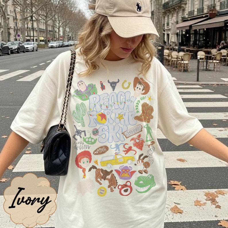 Pode incluir: Camiseta cor marfim com um design gr&aacute;fico colorido com personagens de desenhos animados e o texto "REACH FOR THE SKY". A camiseta &eacute; combinada com um bon&eacute; de beisebol bege e uma bolsa preta com al&ccedil;a de corrente. A camiseta tem um ajuste folgado.