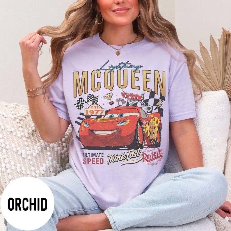Op de afbeelding: Orchidee-kleurig T-shirt met een afbeelding van Lightning McQueen uit de film Cars. Het ontwerp bevat de tekst "Lightning McQueen" en "Cars", samen met racevlaggen en het jaar "1977".