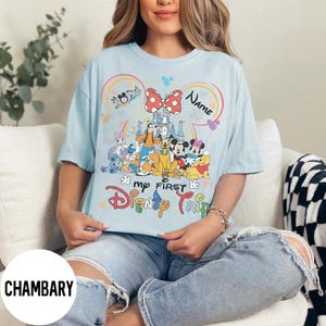 May include: Light blue t-shirt featuring a colorful Disney graphic. The design includes Mickey and Minnie Mouse, Winnie the Pooh, and other characters. Text on the shirt reads "My First Disney Trip" and "Name".