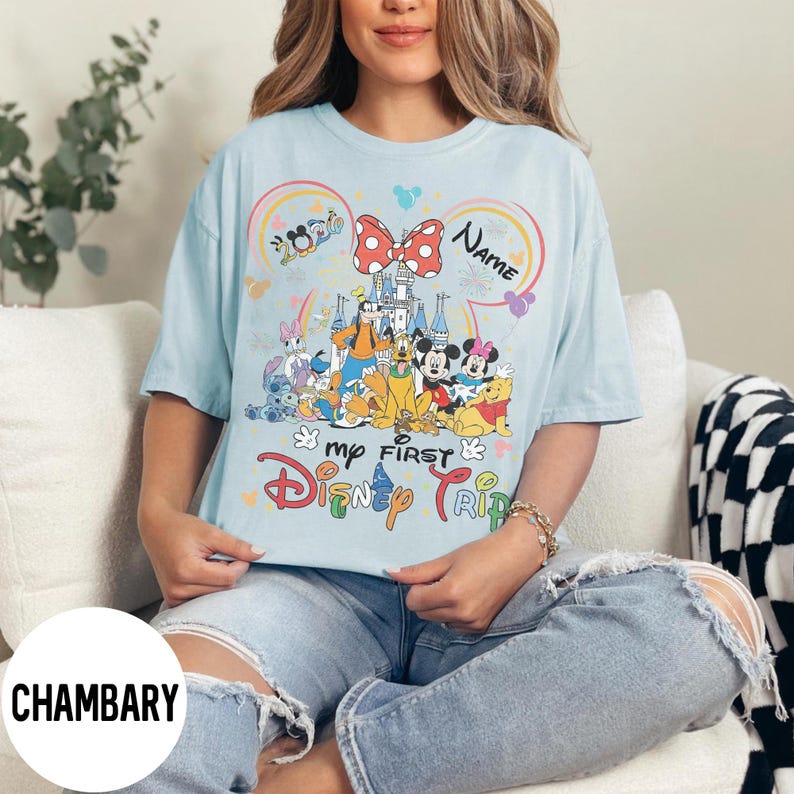 May include: Light blue t-shirt featuring a colorful Disney graphic. The design includes Mickey and Minnie Mouse, Winnie the Pooh, and other characters. Text on the shirt reads "My First Disney Trip" and "Name".