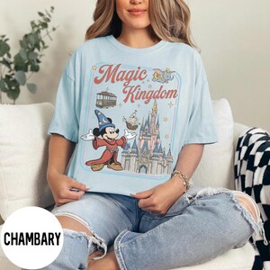 May include: Pale blue t-shirt with a graphic featuring Mickey Mouse in a sorcerer's outfit, a castle, and the words "Magic Kingdom." The design includes a trolley car, a ship, and Dumbo the elephant. The shirt has a relaxed fit.