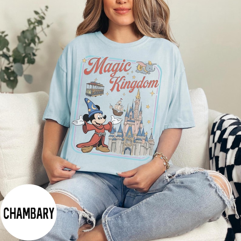 May include: Pale blue t-shirt with a graphic featuring Mickey Mouse in a sorcerer's outfit, a castle, and the words "Magic Kingdom." The design includes a trolley car, a ship, and Dumbo the elephant. The shirt has a relaxed fit.