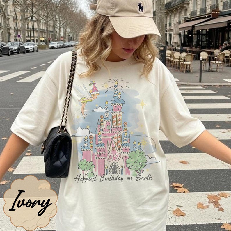 Puede incluir: Camiseta de color marfil con un gr&aacute;fico de castillo colorido y el texto "Happiest Birthday on Earth". El dise&ntilde;o incluye un hada y velas. La camiseta se combina con una gorra de b&eacute;isbol beige y un bolso negro.