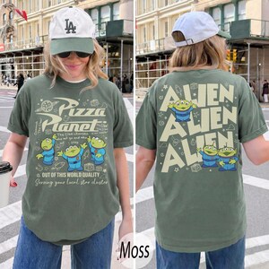 K&ouml;nnte beinhalten: Moosgr&uuml;nes T-Shirt mit dem Pizza Planet-Logo und Alien-Charakteren. Auf der Vorderseite des Shirts befinden sich das Pizza Planet-Logo und Aliens. Auf der R&uuml;ckseite des Shirts steht das Wort "ALIEN" dreimal wiederholt mit Alien-Charakteren.