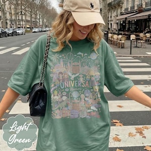 May include: Light green t-shirt with a colourful graphic featuring the Universal Studios logo and cartoon characters. The shirt has short sleeves and a relaxed fit. A beige cap and a black purse are also visible.