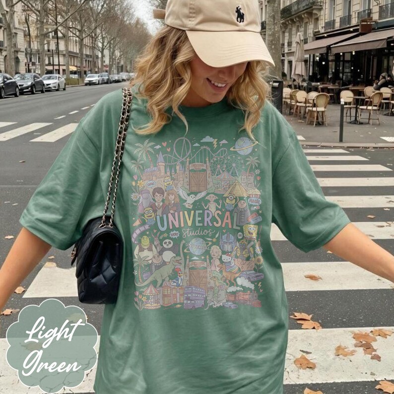 May include: Light green t-shirt with a colourful graphic featuring the Universal Studios logo and cartoon characters. The shirt has short sleeves and a relaxed fit. A beige cap and a black purse are also visible.