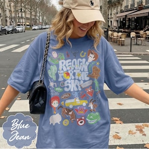 Pode incluir: Camiseta azul jeans com um gr&aacute;fico colorido com personagens e o texto "REACH FOR THE SKY". O design inclui um cacto, botas de cowboy e um caminh&atilde;o amarelo. Uma bolsa preta com al&ccedil;a de corrente &eacute; vis&iacute;vel.