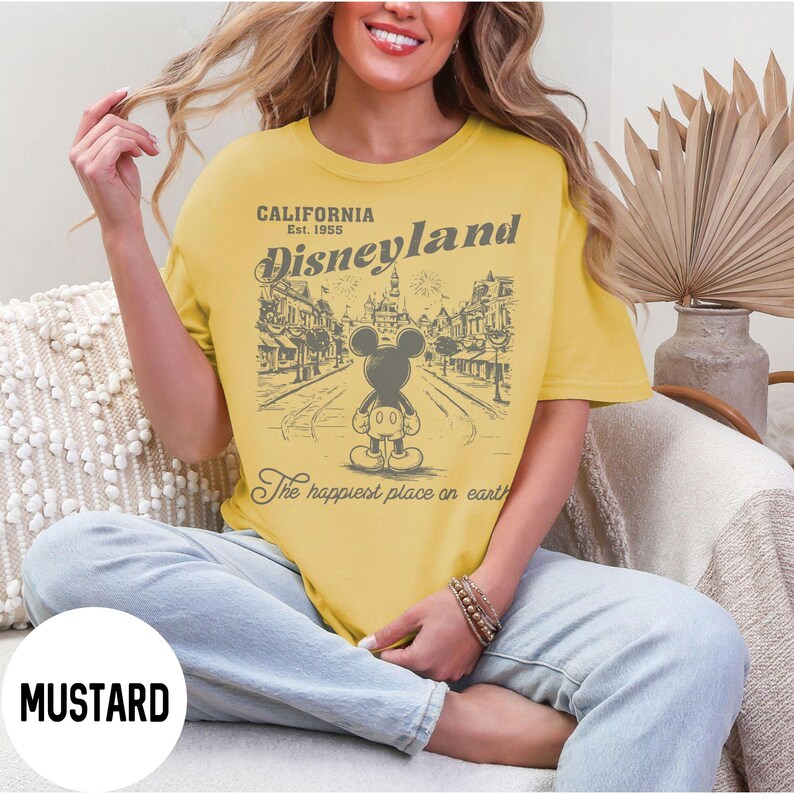 Może przedstawiać: Musztardowa koszulka z grafiką Disneyland. Projekt zawiera napis "CALIFORNIA Est. 1955 Disneyland" nad ilustracją Myszki Miki. Poniżej znajduje się fraza "The happiest place on earth". Koszulka jest w kolorze musztardowym.