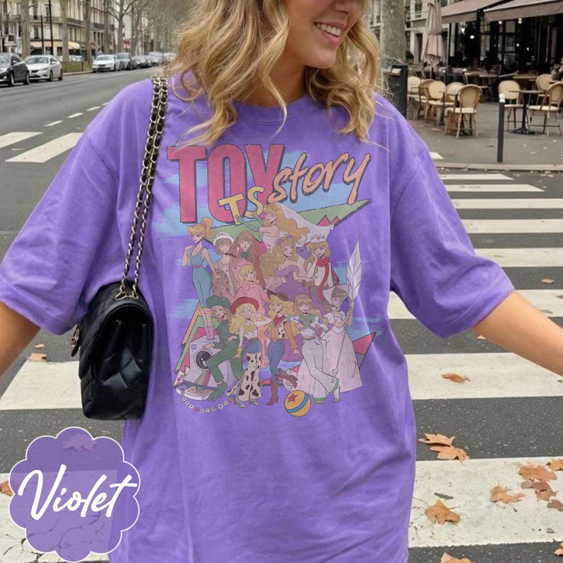 May include: A violet t-shirt featuring a graphic print with the text Toy Story. The design displays a colorful illustration of various animated characters in a group, including figures in diverse outfits, a small ball, and a camera on a light blue background.