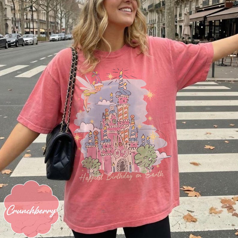 Puede incluir: Camiseta de color coral con una ilustraci&oacute;n de castillo caprichosa, un hada y el texto "Happiest Birthday on Earth". El dise&ntilde;o incluye velas, estrellas y &aacute;rboles. La camiseta tiene mangas cortas y un corte relajado.