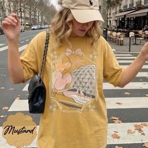 May include: Mustard-colored t-shirt featuring a graphic design with a pink bow, Mickey Mouse ears, a geometric sphere, and a monorail. The word "Mustard" is written in cursive at the bottom of the shirt.