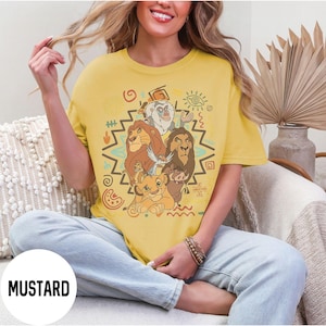 Puede incluir: Camiseta de color mostaza con un gr&aacute;fico de personajes de El Rey Le&oacute;n. El dise&ntilde;o incluye a Simba, Mufasa, Scar, Tim&oacute;n, Pumba y Rafiki, con patrones de inspiraci&oacute;n tribal. La palabra "MUSTARD" est&aacute; impresa en la parte inferior izquierda.