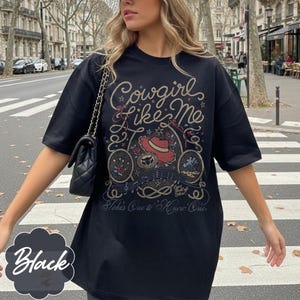 May include: A black short-sleeve t-shirt featuring a graphic print with rope-style lettering that reads Cowgirl Like Me and Takes One to Know One. The design includes a red cowboy hat, a horseshoe, a cactus, and musical notes in a western style.