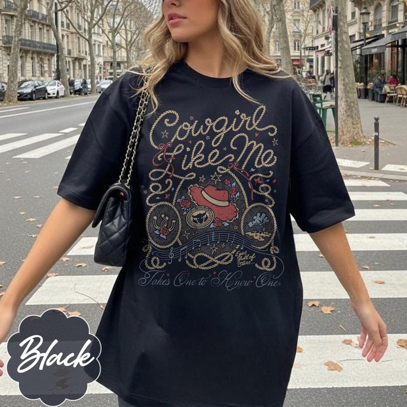 May include: A black short-sleeve t-shirt featuring a graphic print with rope-style lettering that reads Cowgirl Like Me and Takes One to Know One. The design includes a red cowboy hat, a horseshoe, a cactus, and musical notes in a western style.