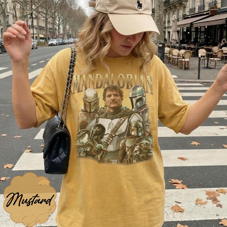 Pu&ograve; includere: Una t-shirt a maniche corte color giallo senape con una stampa grafica dei personaggi di The Mandalorian. Il design include il testo "MANDALORIAN" in un carattere oro metallizzato sopra una figura centrale circondata da personaggi in armatura.