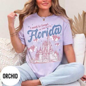 May include: Orchid-colored t-shirt with the text "I need to forget so take me to Florida" in blue script. The design includes a castle illustration, fireworks, and Mickey Mouse balloon graphics.