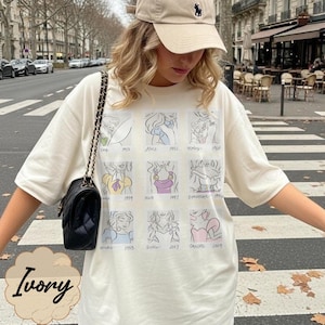May include: Ivory-colored t-shirt featuring a grid of cartoon sketches of animated characters. The shirt has a crew neck and short sleeves. The word "Ivory" is printed at the bottom of the shirt.