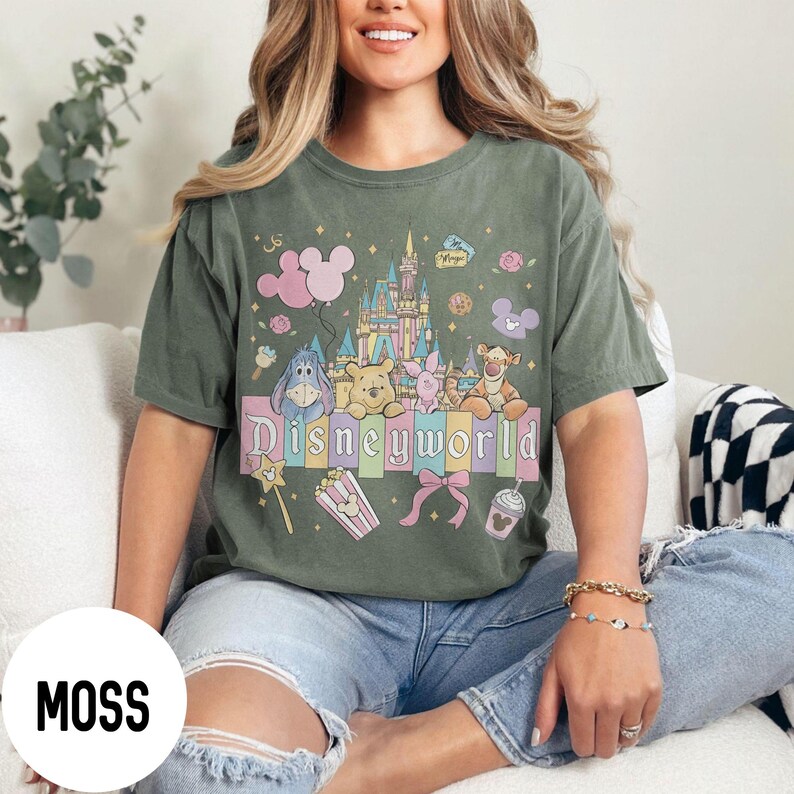 May include: Moss green t-shirt featuring a colorful Disney World graphic. The design includes a castle, balloons, and various Disney characters like Winnie the Pooh and Piglet. The word "Disneyworld" is prominently displayed.