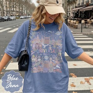 May include: Blue jean coloured t-shirt featuring a colourful graphic of Universal Studios attractions. The graphic includes the text "UNIVERSAL Studios" and cartoon characters. The shirt has a relaxed fit.