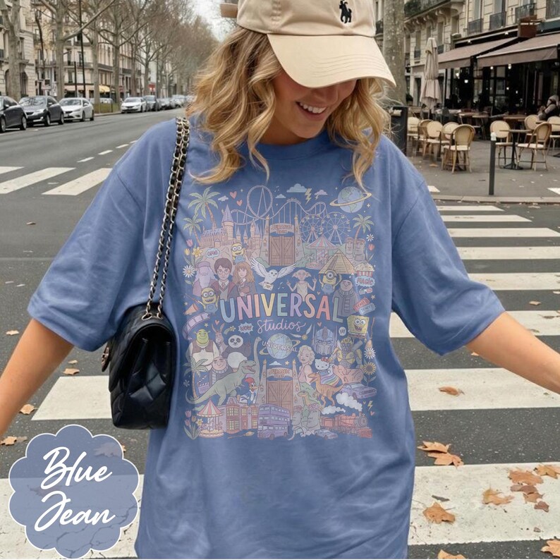 May include: Blue jean coloured t-shirt featuring a colourful graphic of Universal Studios attractions. The graphic includes the text "UNIVERSAL Studios" and cartoon characters. The shirt has a relaxed fit.