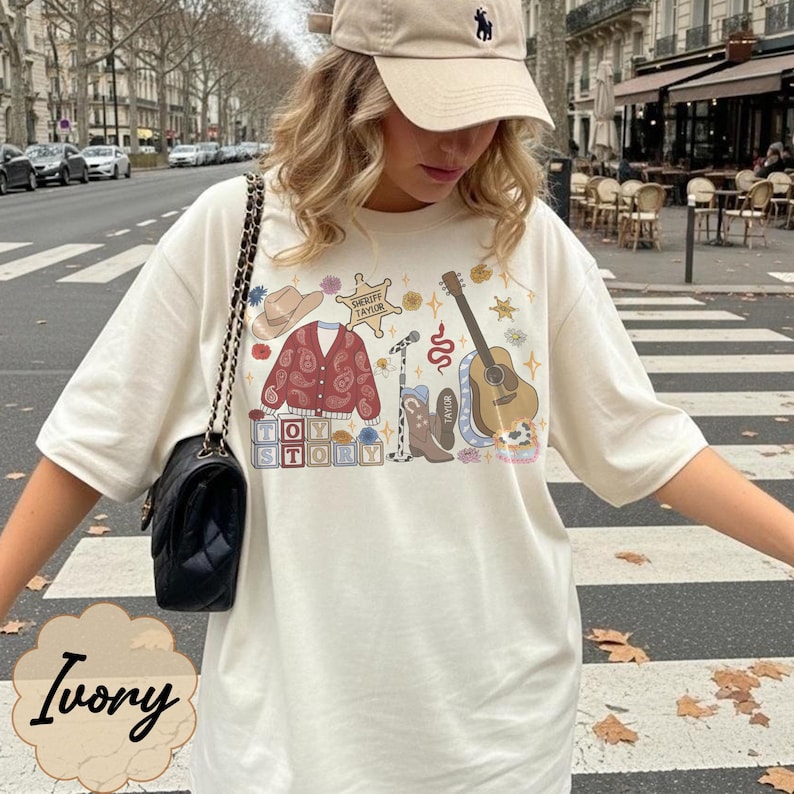 May include: An ivory short-sleeved t-shirt featuring a graphic print of a cowboy hat, a red paisley cardigan, a sheriff badge labeled Sheriff Taylor, a microphone, a brown cowboy boot, a guitar, and blocks spelling out Toy Story.
