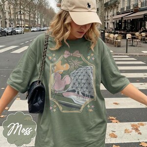 May include: Moss green t-shirt featuring a graphic design with a pink bow, Mickey Mouse, and a geometric sphere. The shirt has short sleeves and a relaxed fit. A beige baseball cap and a black purse with a chain strap are also visible.