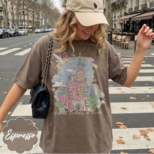 Puede incluir: Camiseta color espresso con una colorida ilustraci&oacute;n de un castillo y el texto "Happiest Birthday on Earth". El dise&ntilde;o incluye un hada y estrellas. La camiseta se lleva con una gorra beige y un bolso negro.