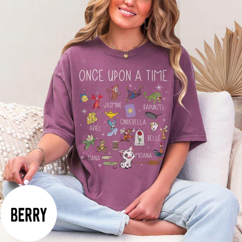 May include: Berry-coloured t-shirt with the text "ONCE UPON A TIME" and various cartoon character illustrations. The shirt features characters like Ariel, Jasmine, Cinderella, and Belle. The word "BERRY" is printed on a white circle.