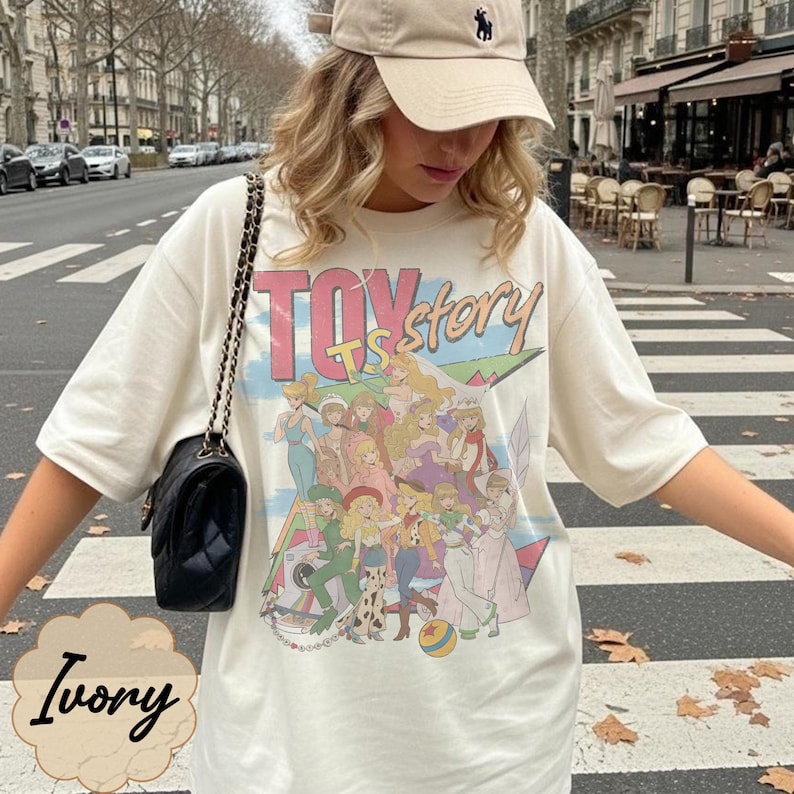 May include: An ivory short-sleeve t-shirt featuring a graphic print with the text "TOY story" and an illustration of various animated characters. The shirt has a crew neckline and a loose fit, paired with a black quilted shoulder bag with a chain strap.