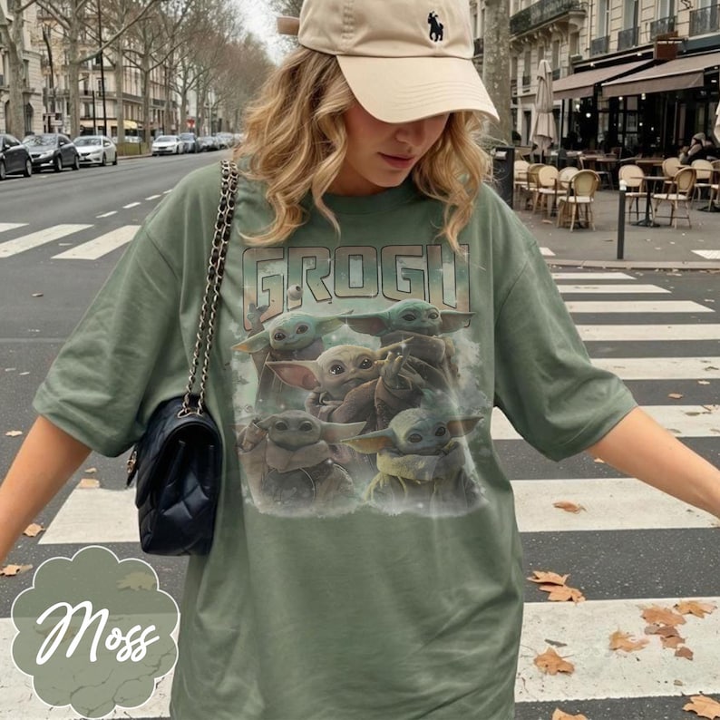 May include: An olive green short-sleeve t-shirt featuring a graphic print of multiple Grogu characters and the text GROGU in a distressed font. The shirt is paired with a beige baseball cap and a black quilted shoulder bag with a chain strap.