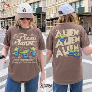 K&ouml;nnte beinhalten: Espressofarbenes T-Shirt mit dem Pizza Planet-Logo und Alien-Charakteren. Auf der Vorderseite des Shirts steht "Pizza Planet" und "Out of this world quality". Auf der R&uuml;ckseite des Shirts steht "Alien" und Alien-Charaktere.