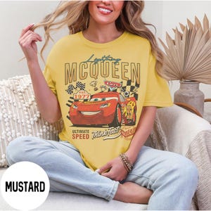Op de afbeelding: Mosterdkleurig T-shirt met Lightning McQueen uit de film Cars. De afbeelding bevat de tekst "Lightning McQueen", "Ultimate Speed" en "Think Fast". Het ontwerp toont McQueen met racevlaggen en het jaar "1977".