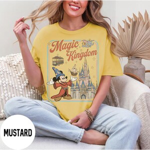 May include: Mustard-coloured t-shirt with a retro graphic of Mickey Mouse as a sorcerer, the Magic Kingdom logo, and a castle. The design includes a tram, ship, and Dumbo. The shirt has a relaxed fit.