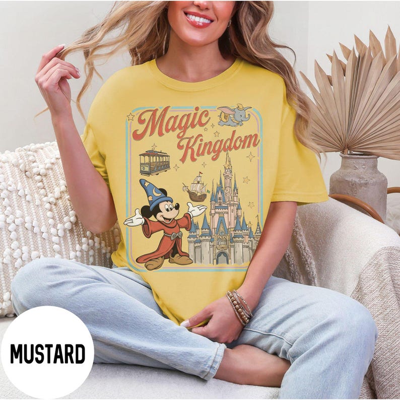 May include: Mustard-coloured t-shirt with a retro graphic of Mickey Mouse as a sorcerer, the Magic Kingdom logo, and a castle. The design includes a tram, ship, and Dumbo. The shirt has a relaxed fit.