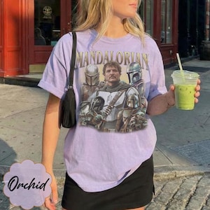 Pu&ograve; includere: Una t-shirt a maniche corte color viola chiaro con una stampa grafica dei personaggi di The Mandalorian. Il design include il testo "MANDALORIAN" in un carattere dorato sopra una figura centrale circondata da personaggi corazzati e una piccola creatura verde.