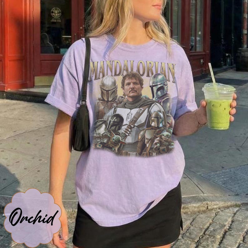 Pu&ograve; includere: Una t-shirt a maniche corte color viola chiaro con una stampa grafica dei personaggi di The Mandalorian. Il design include il testo "MANDALORIAN" in un carattere dorato sopra una figura centrale circondata da personaggi corazzati e una piccola creatura verde.