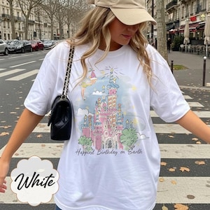 Puede incluir: Camiseta blanca con una ilustraci&oacute;n colorida de un castillo y el texto "Happiest Birthday on Earth". El dise&ntilde;o incluye colores pastel y detalles caprichosos. La palabra "White" tambi&eacute;n est&aacute; impresa en la camiseta.