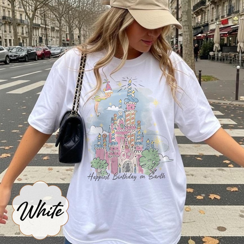 Puede incluir: Camiseta blanca con una ilustraci&oacute;n colorida de un castillo y el texto "Happiest Birthday on Earth". El dise&ntilde;o incluye colores pastel y detalles caprichosos. La palabra "White" tambi&eacute;n est&aacute; impresa en la camiseta.