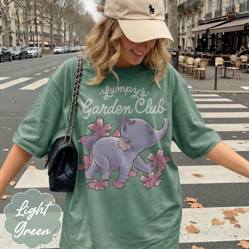 Op de afbeelding: Lichtgroen t-shirt met de tekst "Lumpy's Garden Club" en een cartoon olifantenillustratie. De olifant is paars met roze bloemen. De woorden "Light Green" staan in een lichtgroene bubbel.