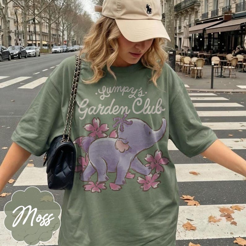 Op de afbeelding: Mosgroen t-shirt met de tekst "Lumpy's Garden Club" en een paarse olifant afbeelding omringd door roze bloemen. Het shirt is een casual stijl met korte mouwen.